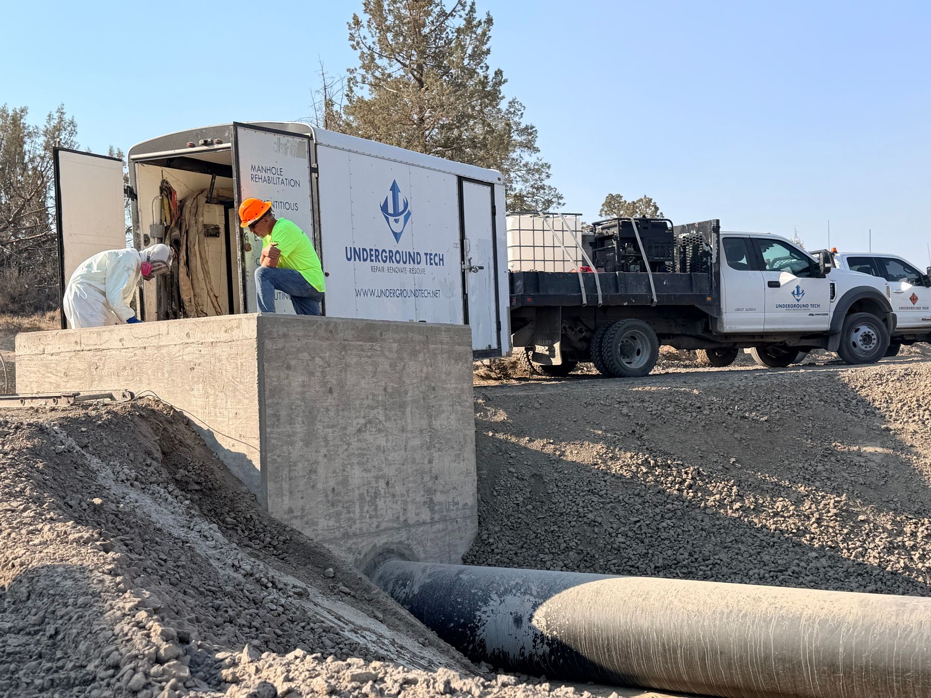 Workers unloading equipment from trucks at a gravel site near a concrete drainage structure.