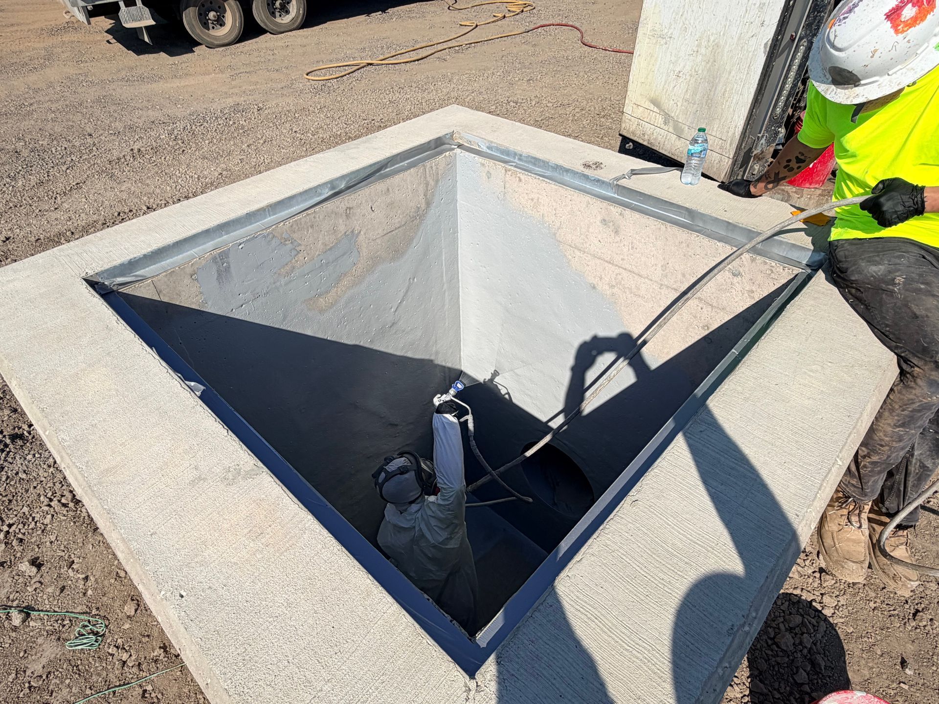 Open square utility pit with concrete walls; worker in safety gear inspecting at right edge.