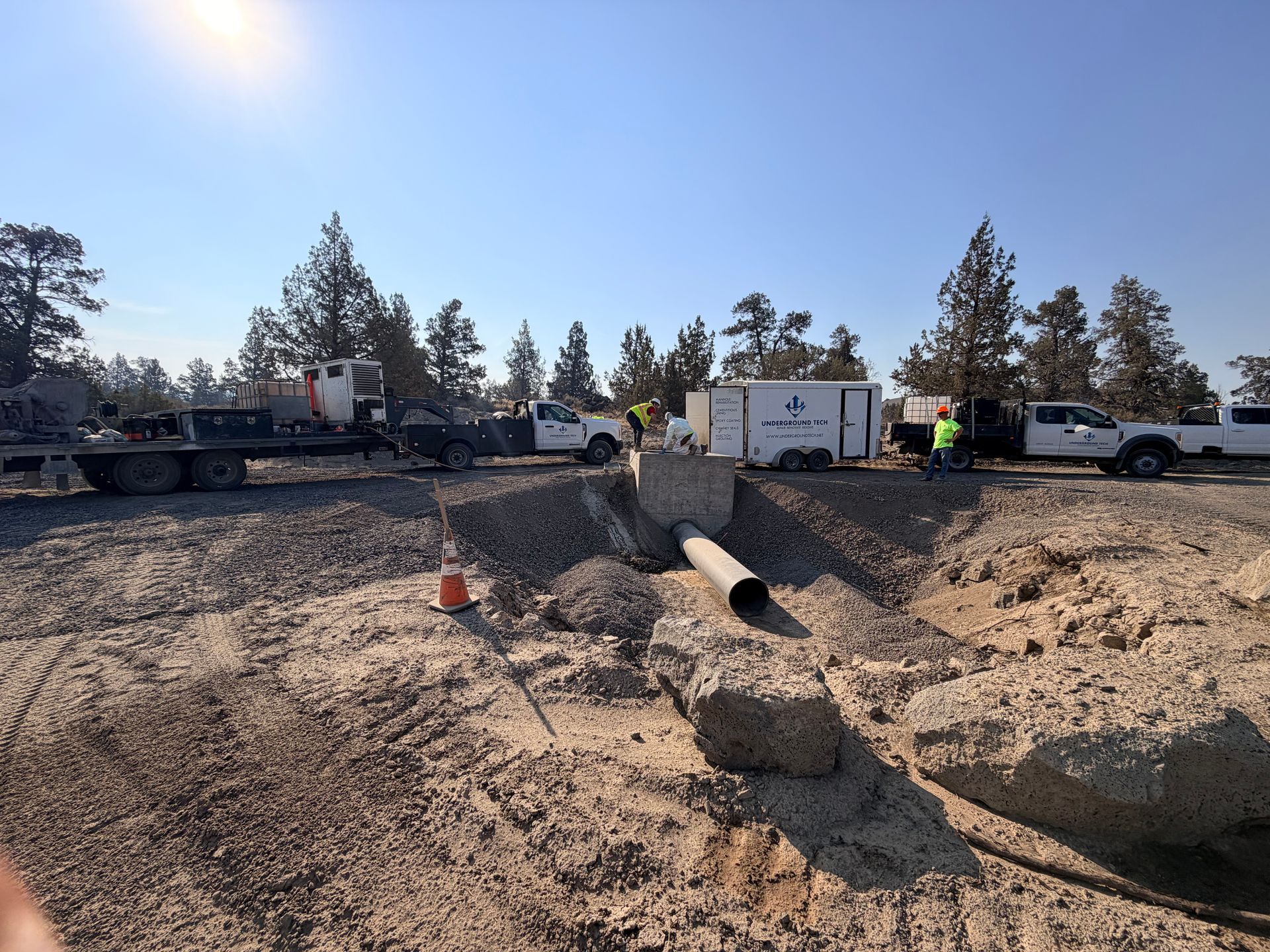 Construction site with dirt trench, exposed pipe, and trucks parked under a clear blue sky