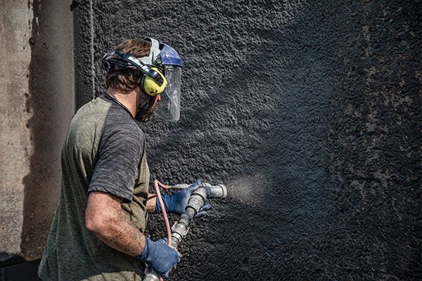 Worker spray-painting a dark wall outdoors, wearing ear protection and gloves