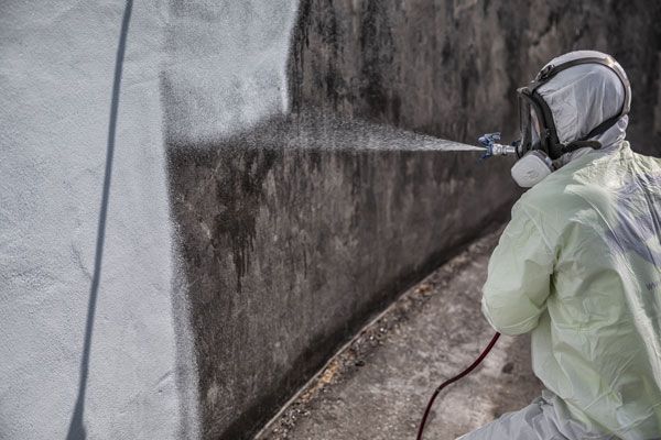 Person in protective suit pressure-washing a dark exterior wall in the rain