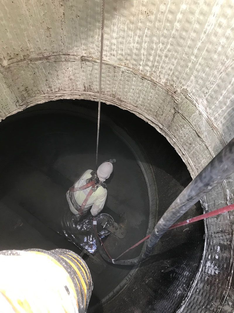 Worker descending into a large cylindrical tank by rope, with water pooled at the bottom.