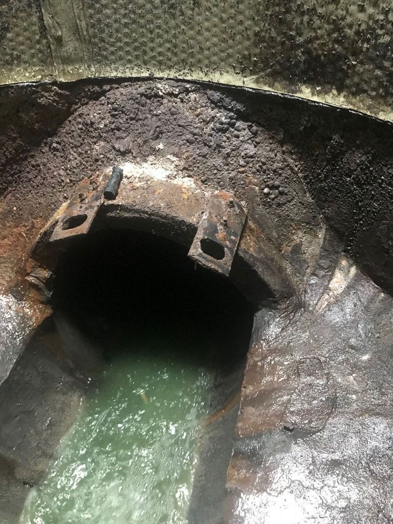A dark concrete drainage tunnel with green water flowing out beneath a metal grate.