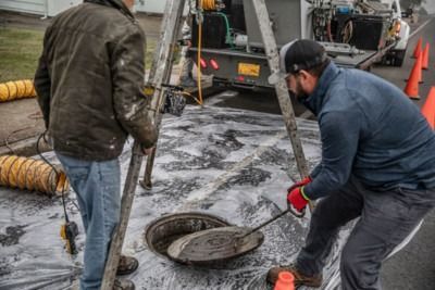 Workers repairing a manhole on a wet street, with traffic cones and tools nearby.