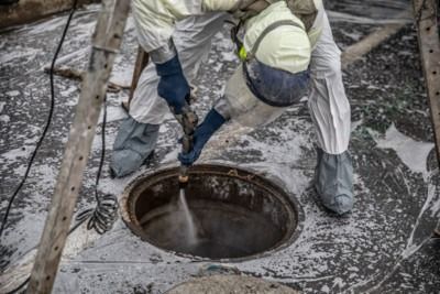 Worker cleaning a manhole with a hose, wearing protective gear and boots on a wet concrete surface