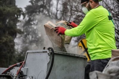 Worker in neon green shirt pouring debris into a dumpster outdoors, with smoke in the background