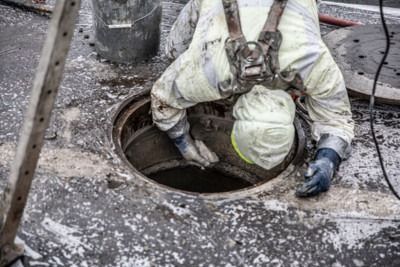 Worker in a harness reaching into a manhole opening on a wet concrete surface.