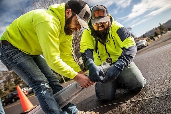 Two workers in neon safety jackets kneel on a road, repairing pavement with tools.