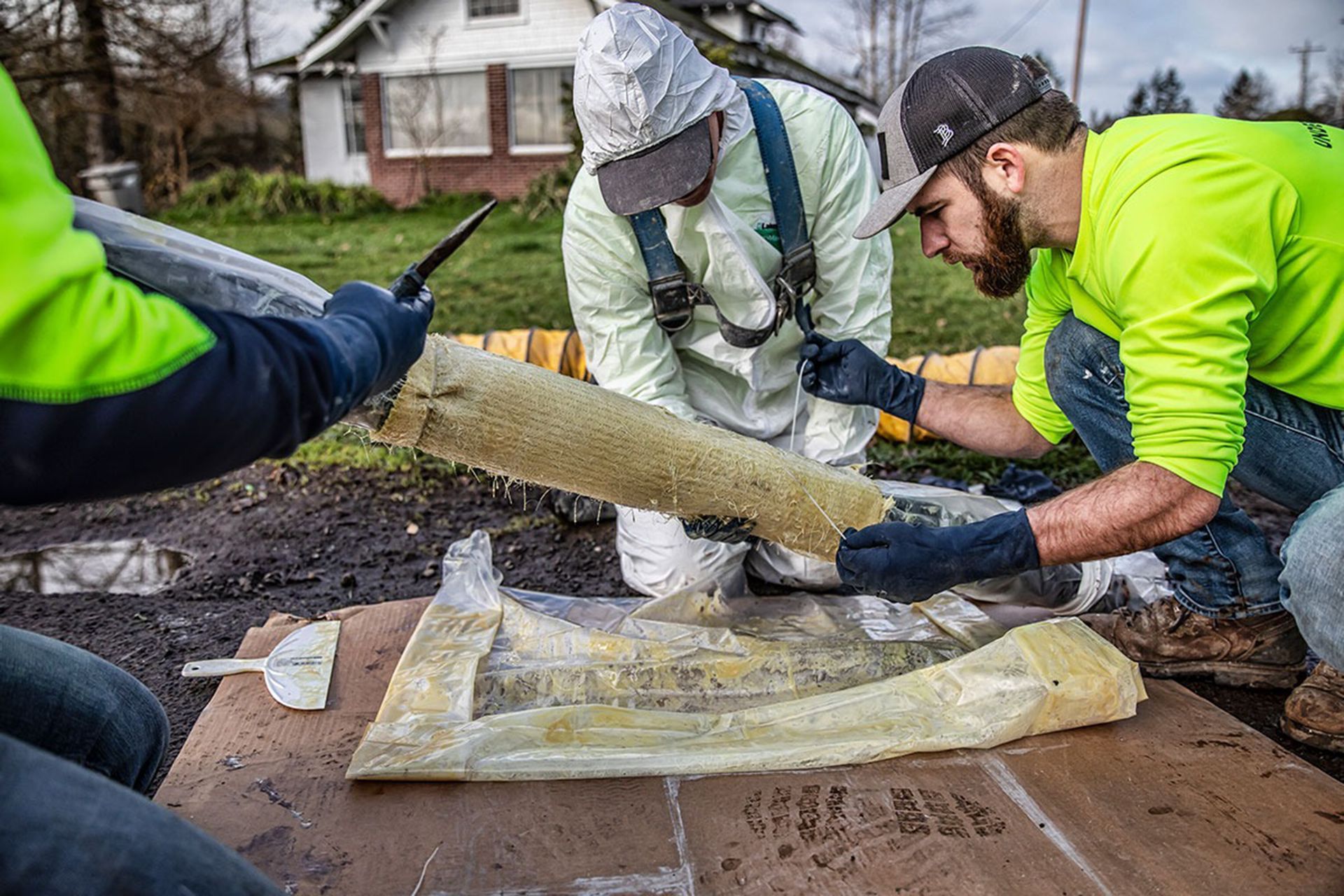 Workers in gloves and rain gear installing a long hose or pipe on a muddy construction site