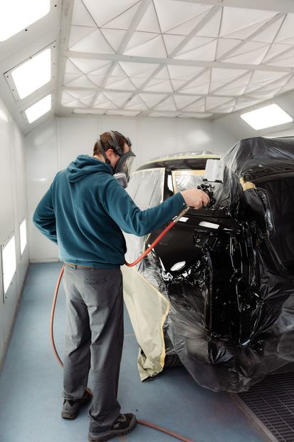 Person spray-painting a black vehicle in a workshop, wearing a respirator and gloves.