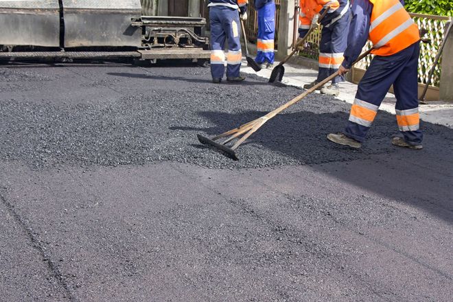 Road workers spreading fresh asphalt on a street with a rake and paver nearby