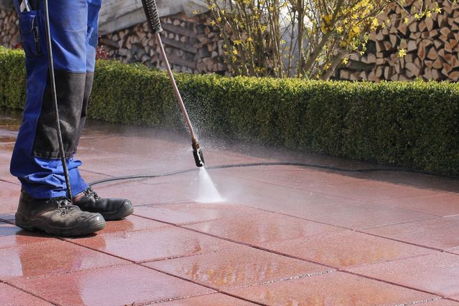 Person pressure-washes a red patio beside stacked firewood and a hedge.