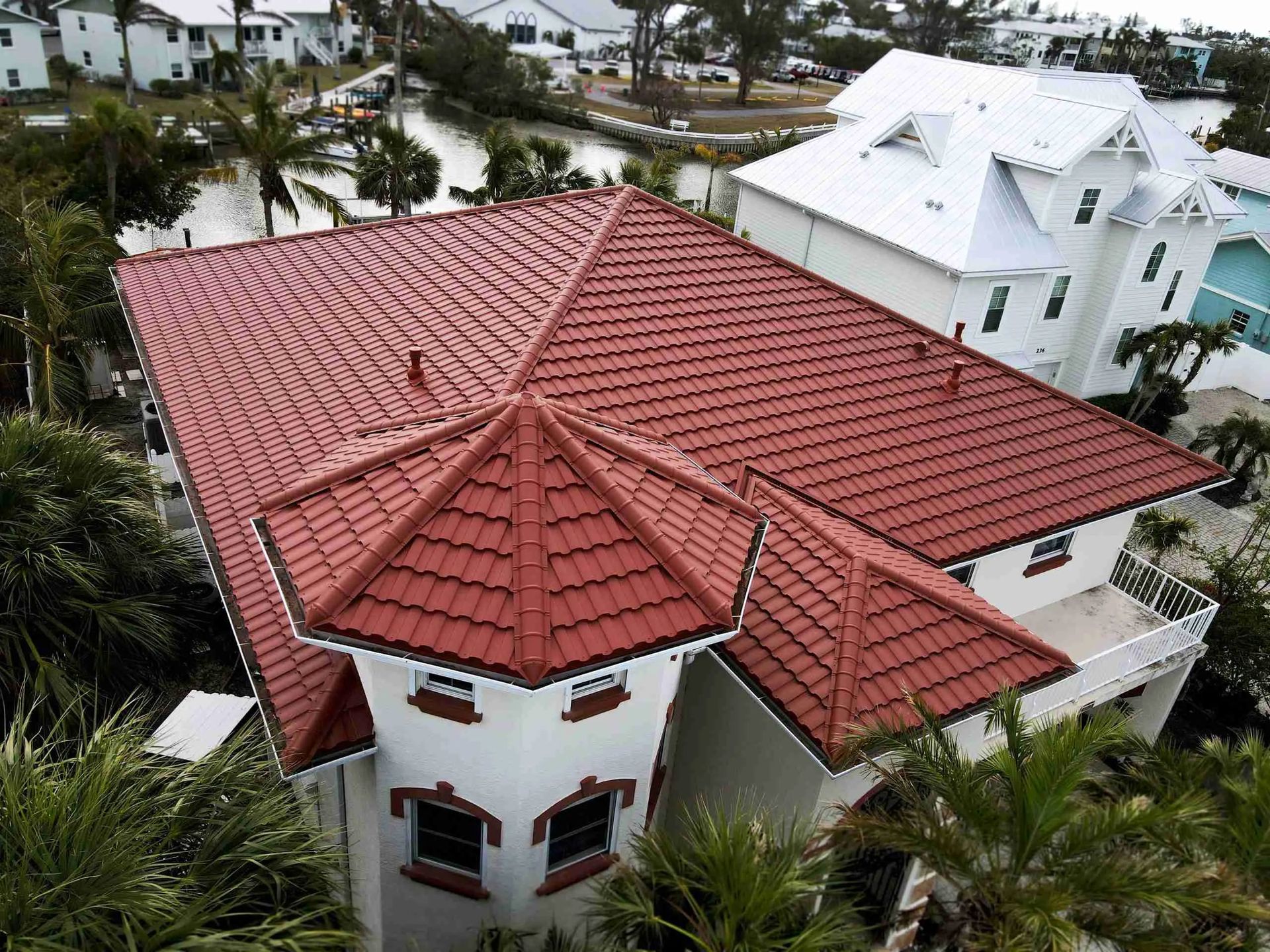 Aerial view of a white house with a red tile roof surrounded by palm trees and nearby homes