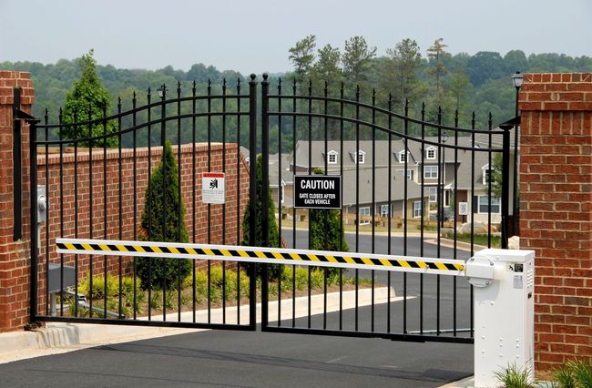 Gated entrance with black wrought-iron gate and yellow-black barrier arm across a driveway