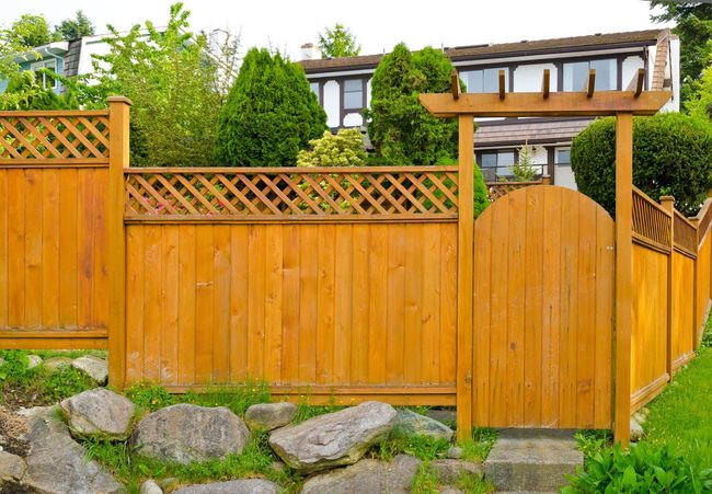 Wooden fence and gate in front of a modern house with greenery and rocks in the yard