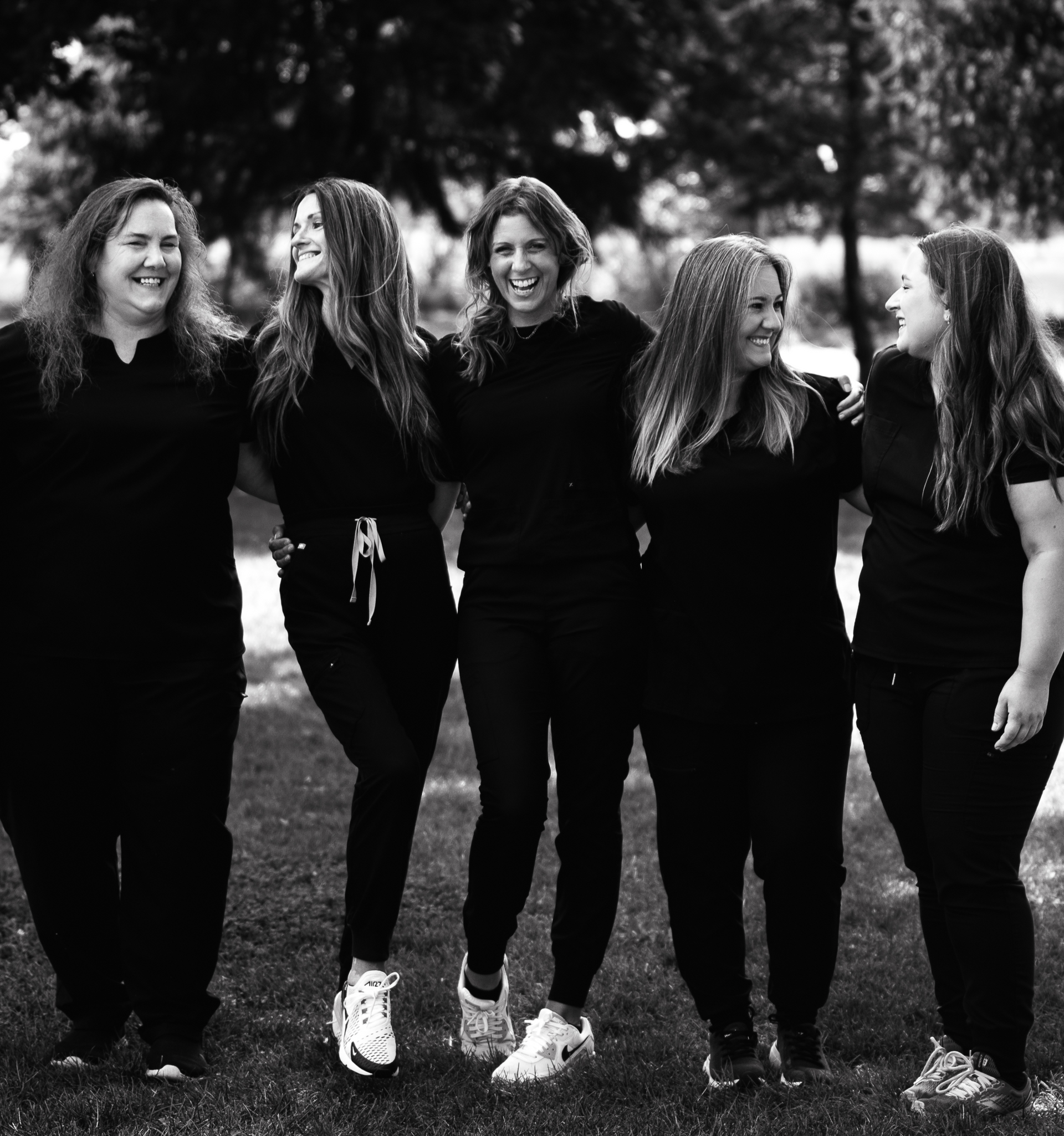 Five people in black clothing stand arm-in-arm outside, smiling.