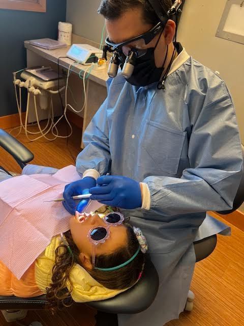 Dentist in blue scrubs examines a child's teeth; child wears protective glasses.