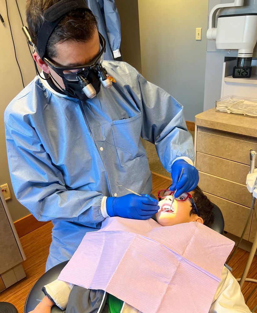 Dentist examining a child's teeth in a dental office. Both are wearing protective gear, and the child is in a dental chair.