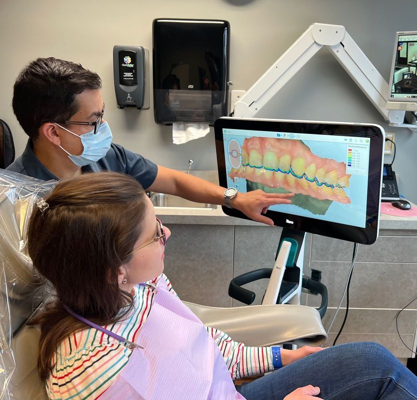 Dentist in mask points to 3D teeth scan on screen for patient in chair. Clinic setting.