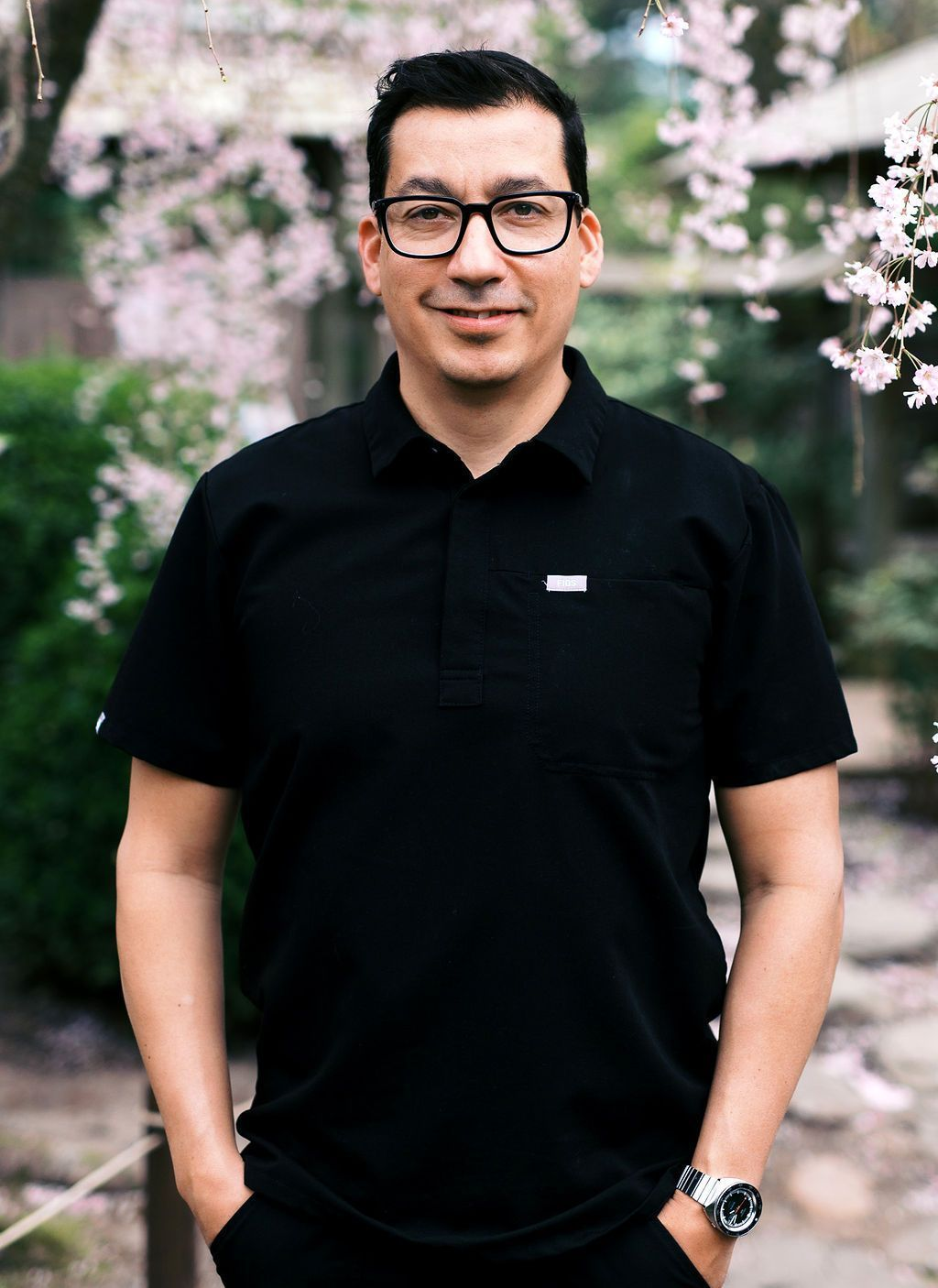 Man in black polo shirt and glasses smiles in a garden with pink flowers.