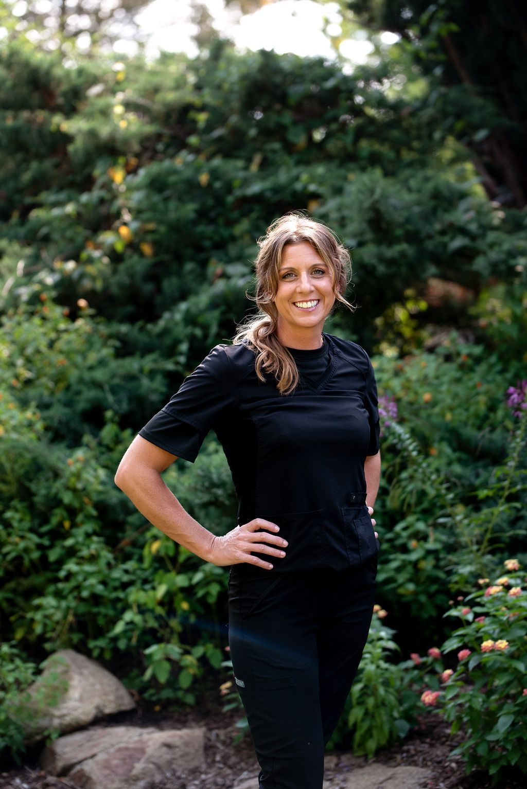 Woman with hand on hip smiles in a garden setting, wearing black.