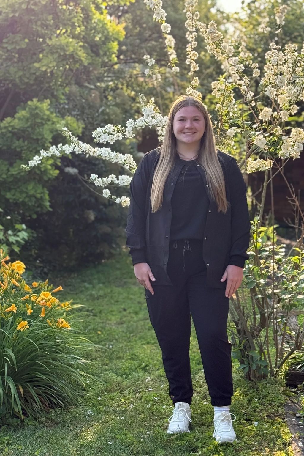 Woman in black scrubs and white shoes standing in a garden, smiling.