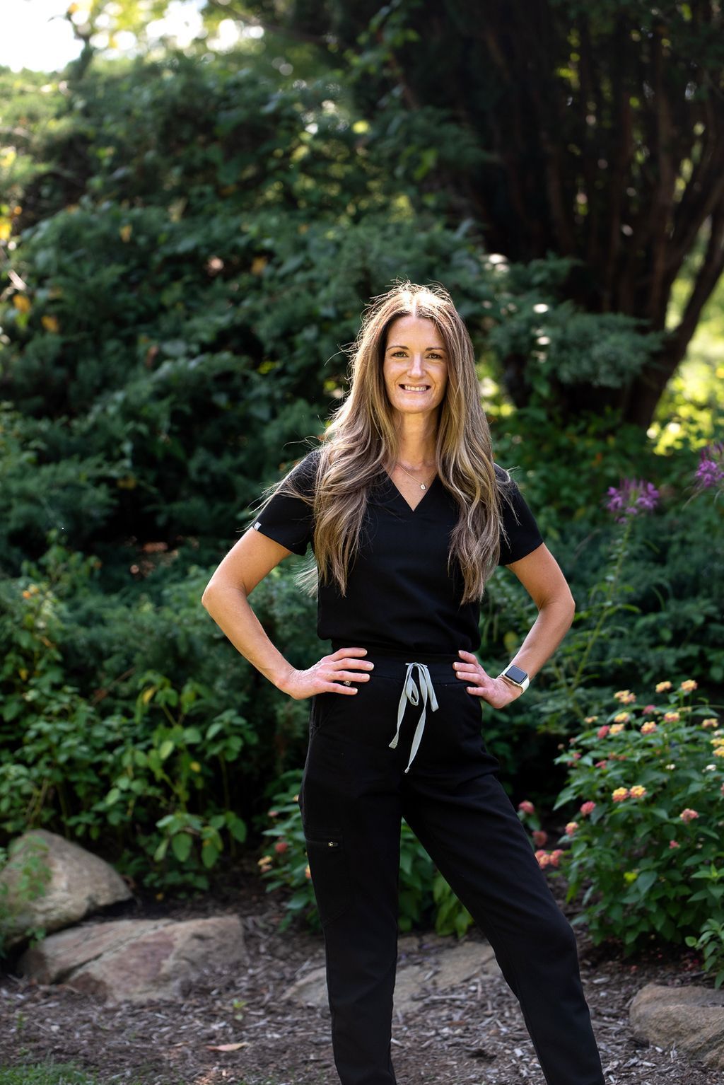 Woman in black scrubs outdoors, hands on hips, smiling, standing by greenery.