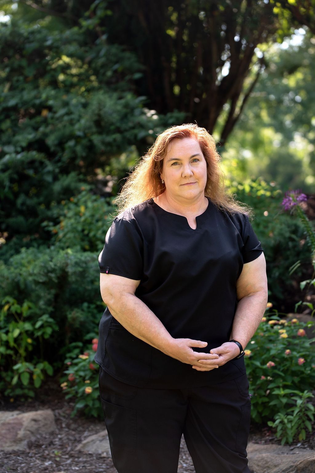 Woman with red hair wearing black scrubs, standing in a garden.