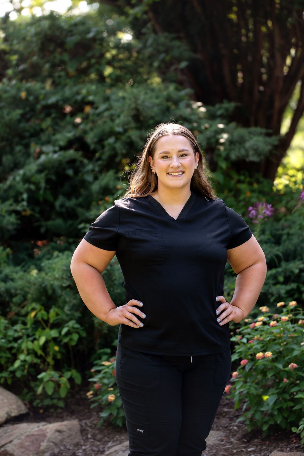 Woman in black scrubs, hands on hips, smiling, standing outside near bushes.
