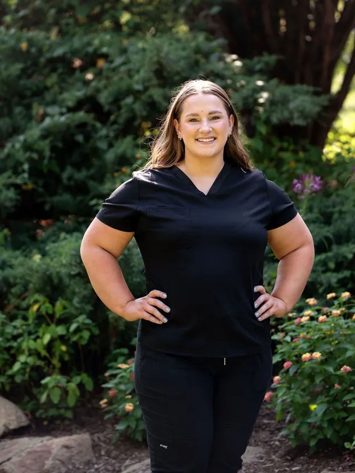 Woman in black scrubs, hands on hips, smiling, standing outside near bushes.