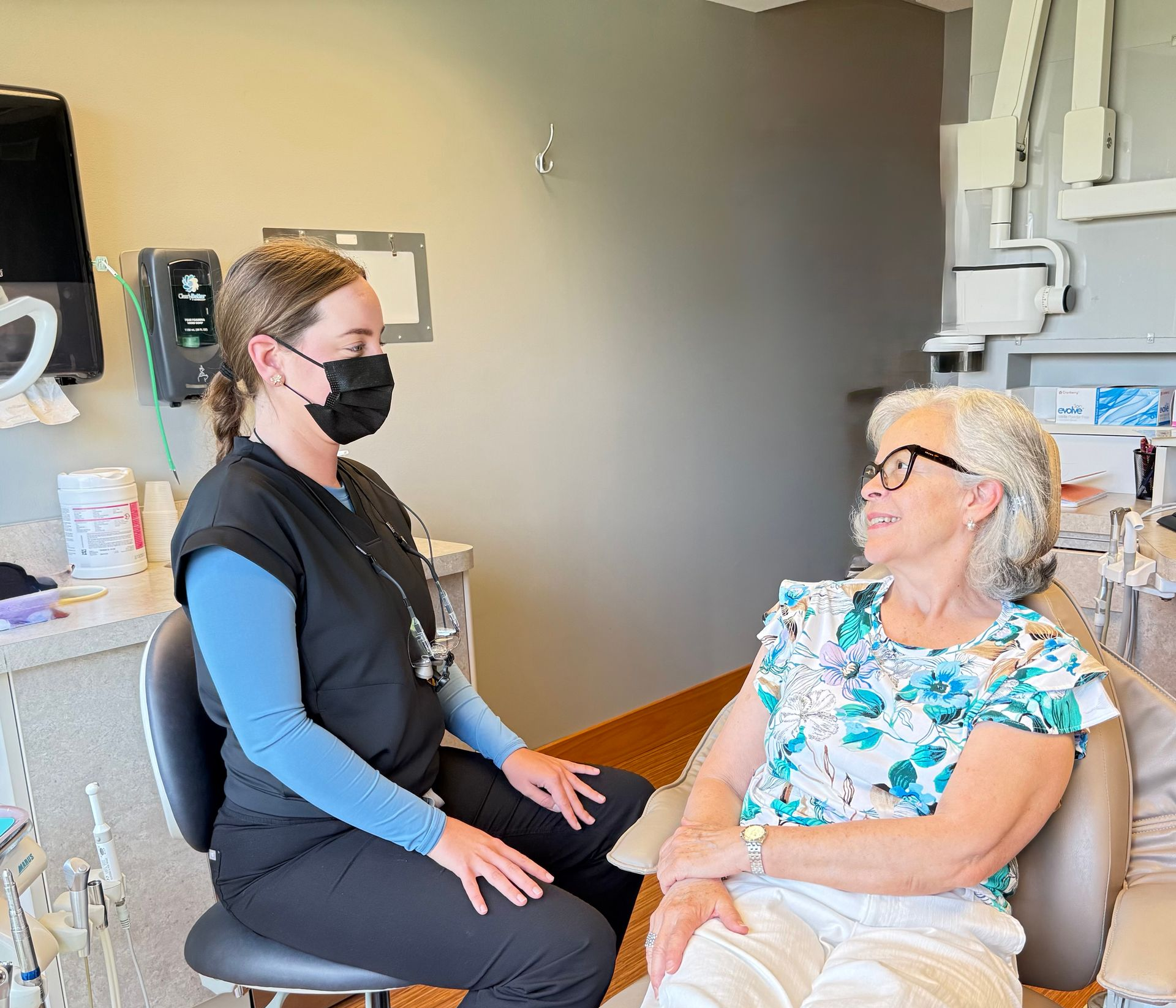 Dentist in black scrubs, mask, consulting with an elderly patient in dental chair, office setting.