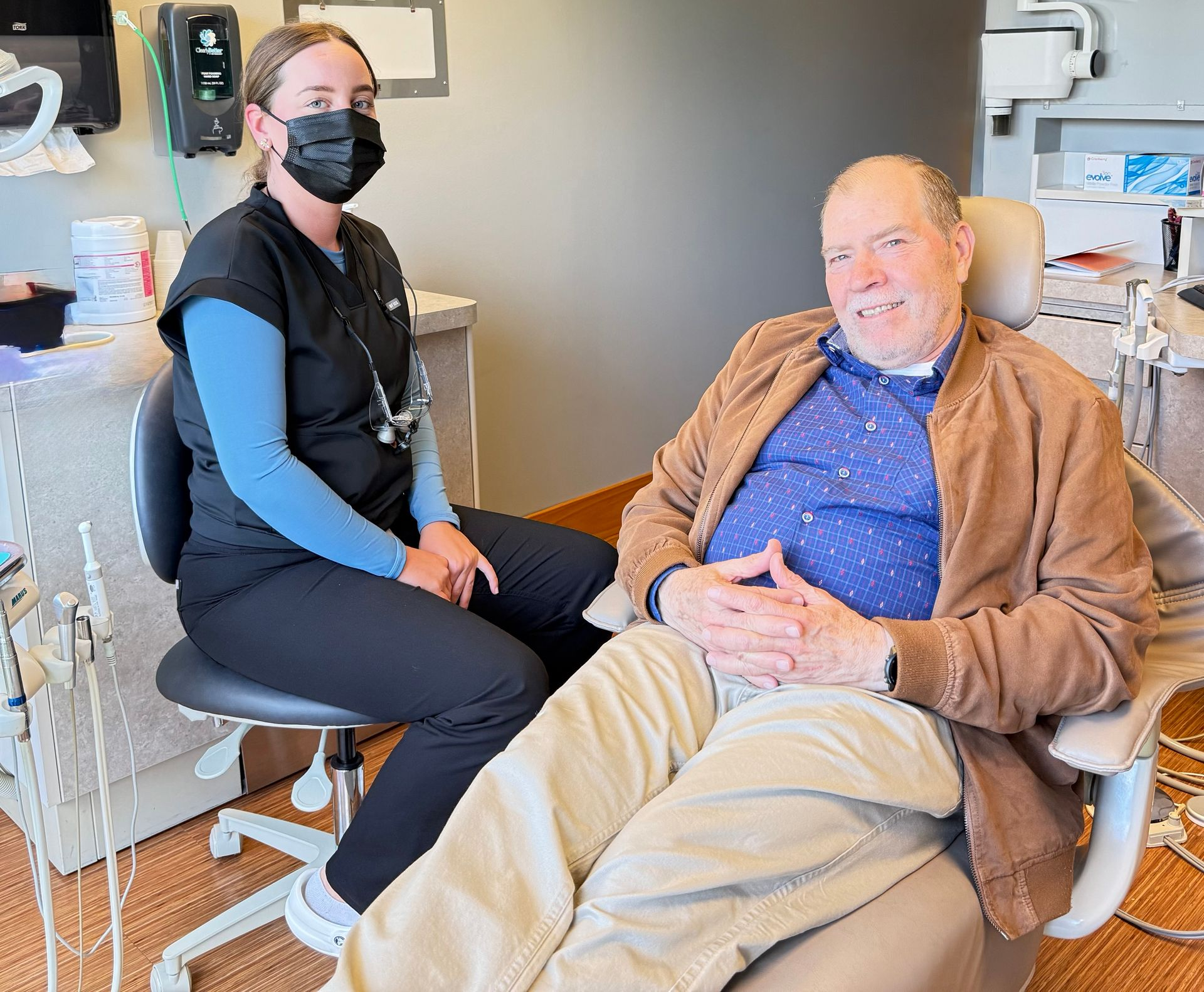 Dental assistant in a mask with a patient in a dental chair, both smiling, in a dental office.