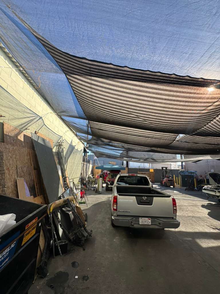 White pickup truck drives under black shade cloth in a narrow outdoor alley with parked items along the sides