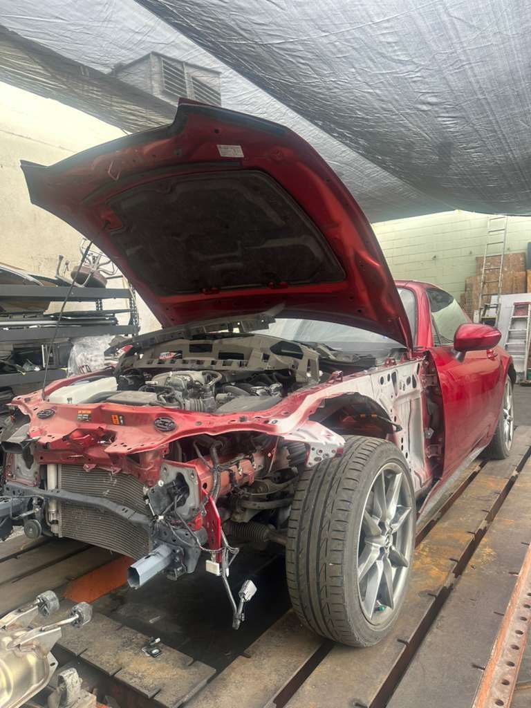 Red car with hood open and front end disassembled on a flatbed tow truck under a tarp shelter.