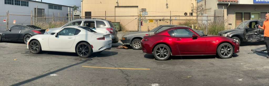 White and red sports cars damaged on a debris-strewn lot in front of an industrial building.