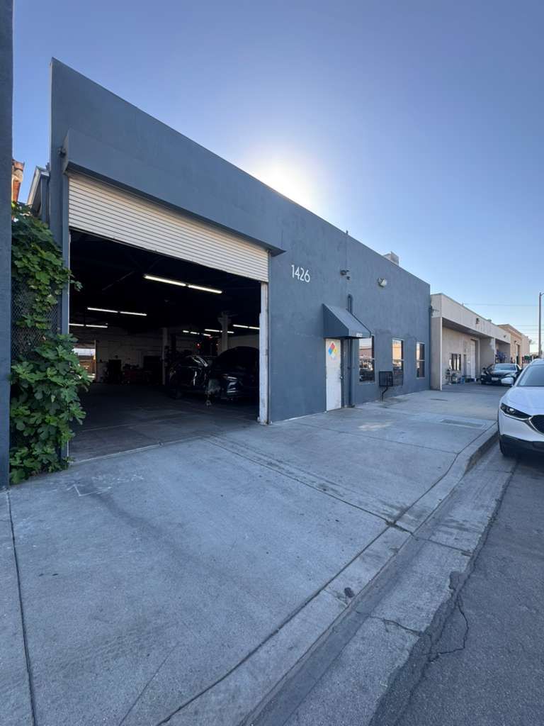 Gray industrial building with open garage bay on a sloped street, parked cars along the curb.