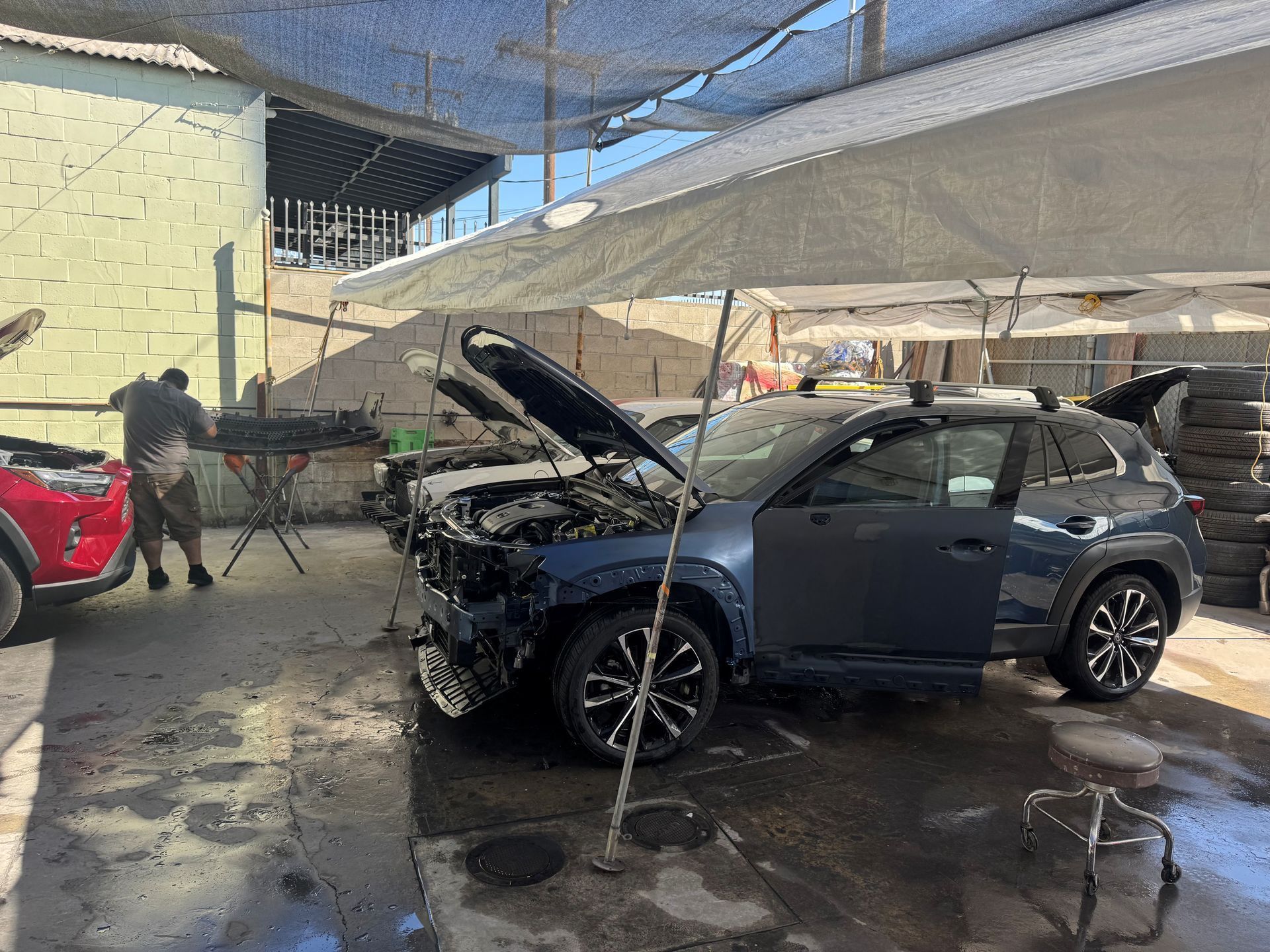 Damaged blue SUV with hood open in a parking lot under shade nets, front end severely crashed.