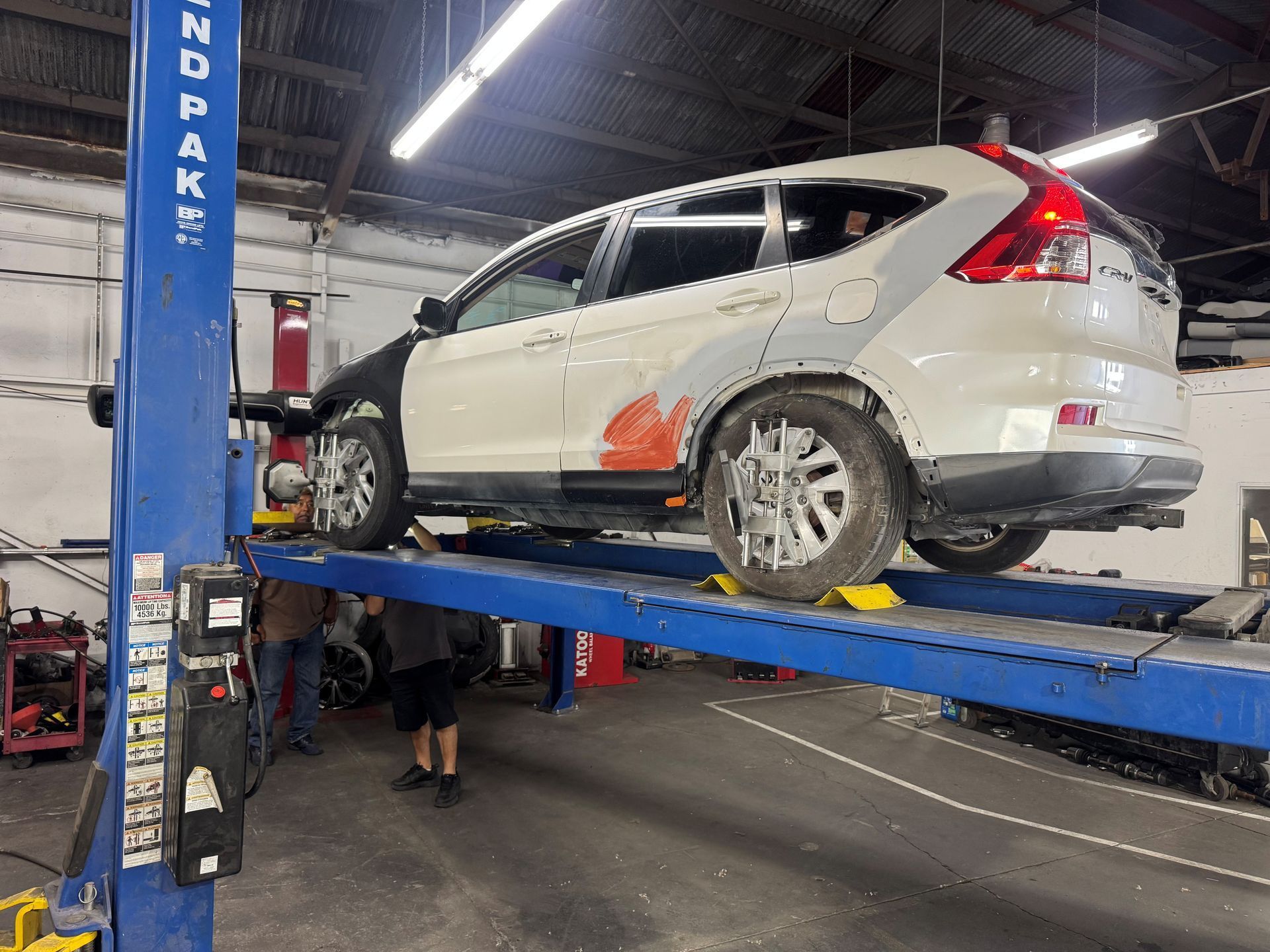 White SUV on a blue automotive lift in a repair shop, with damage visible on the rear side and wheel area.