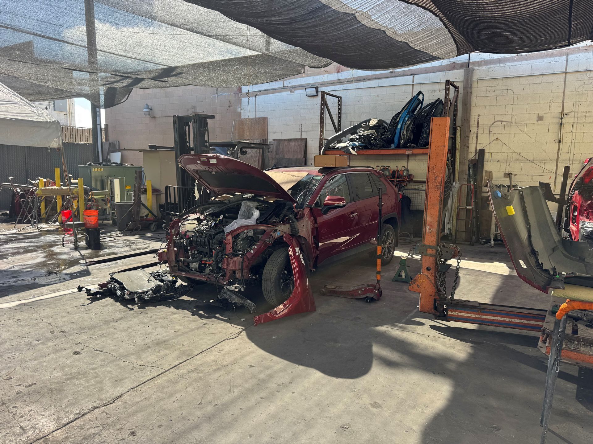 Damaged red car in a garage, front end severely smashed with debris around it.