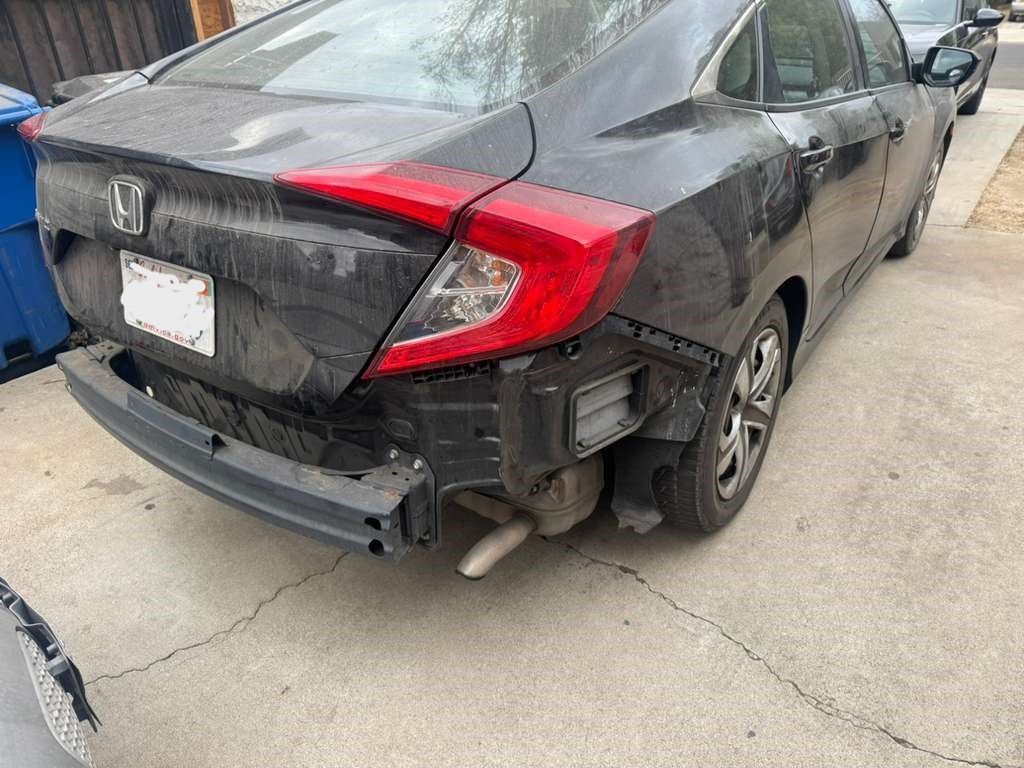 Damaged black Honda sedan with rear bumper and right taillight missing, parked on a concrete driveway.