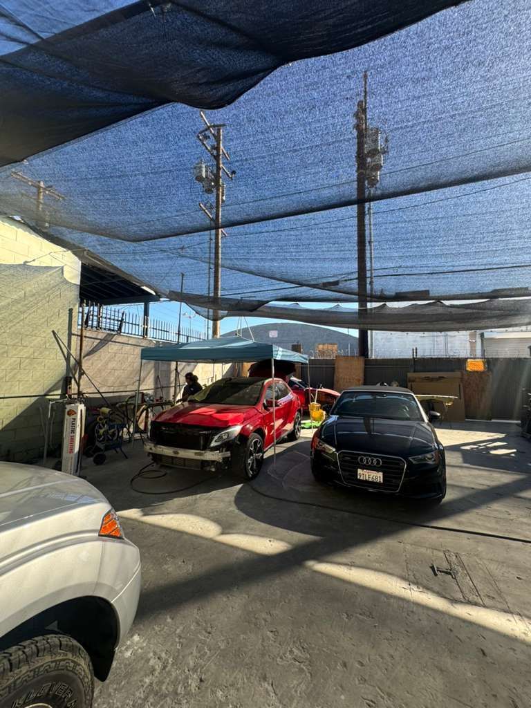 Outdoor car lot with a red race car, black car, and shade netting overhead