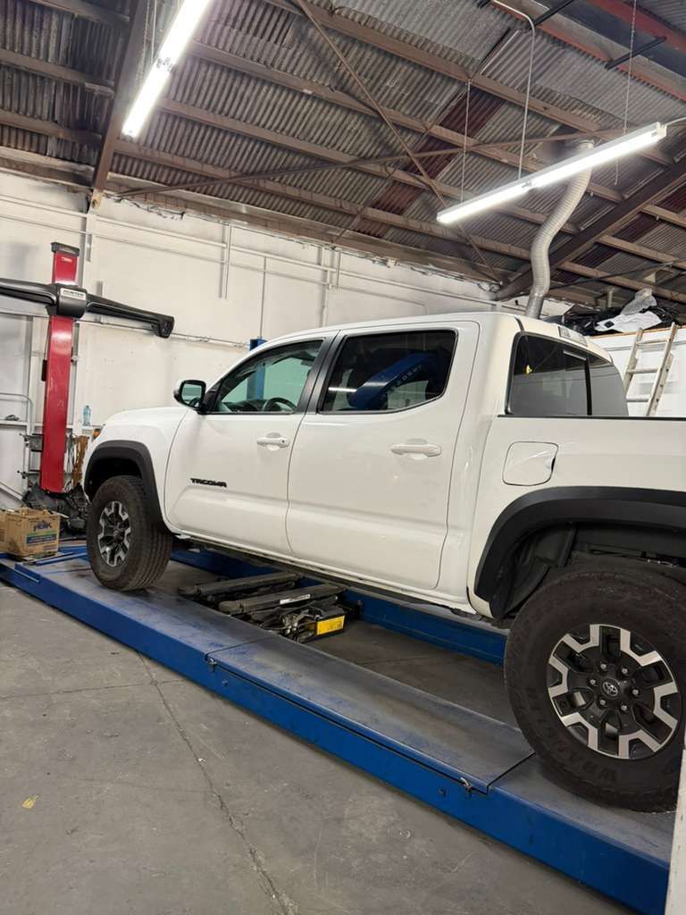 White pickup truck on a blue alignment lift in an auto repair shop with exposed ceiling lights.