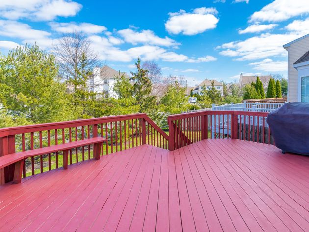Red wooden deck with railing overlooking trees, houses, and a bright blue sky with clouds.