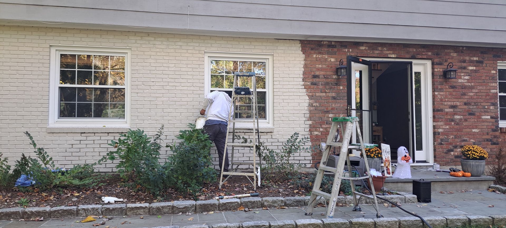 Worker painting the exterior of a house beside a brick wall and window with ladders and supplies nearby
