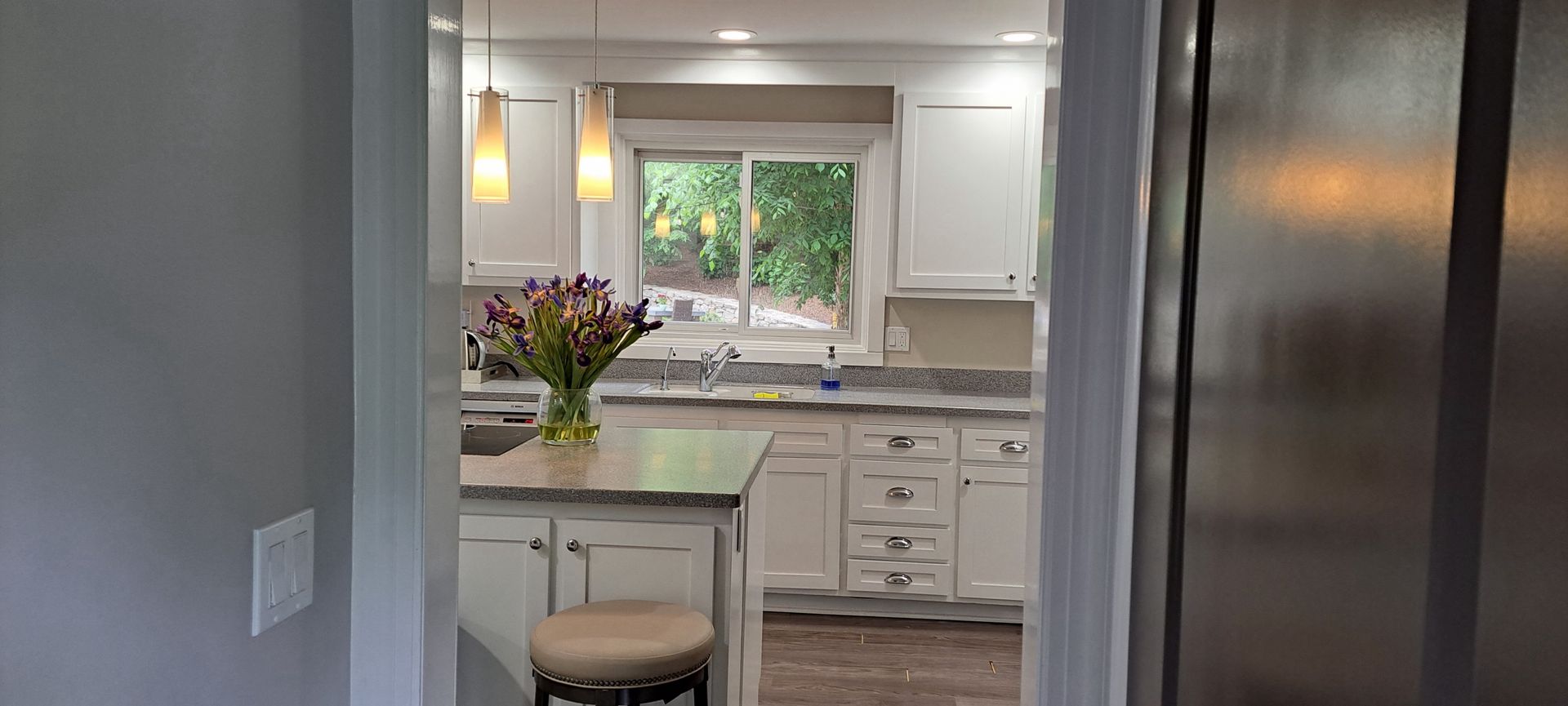Bright white kitchen with island, flowers, stool, and window over the sink.