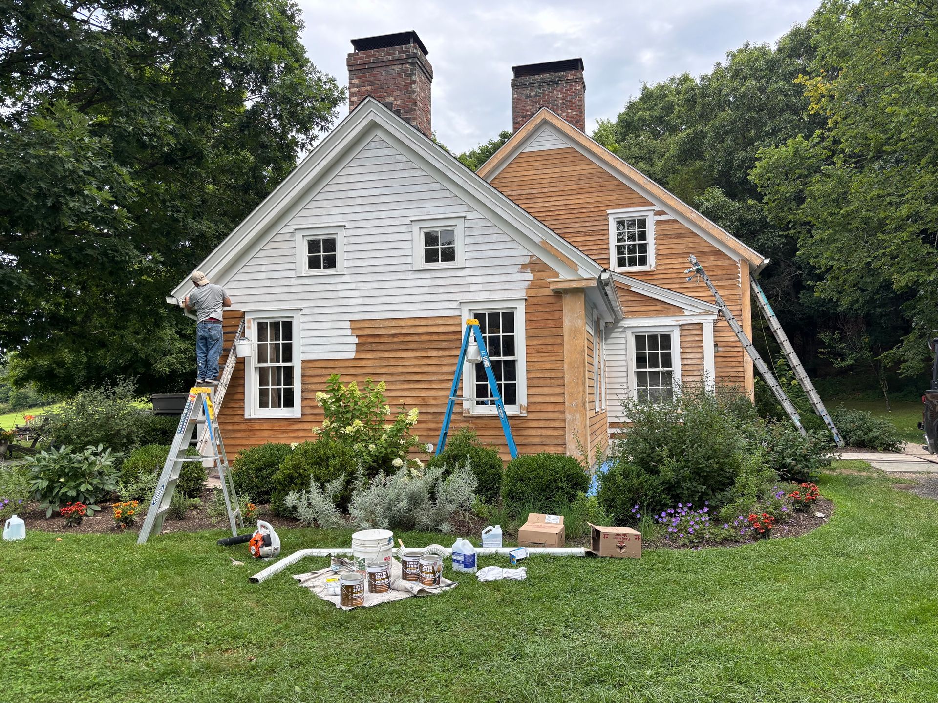 Cottage with white and cedar shingles, ladders, and painting supplies in a green yard with trees behind.