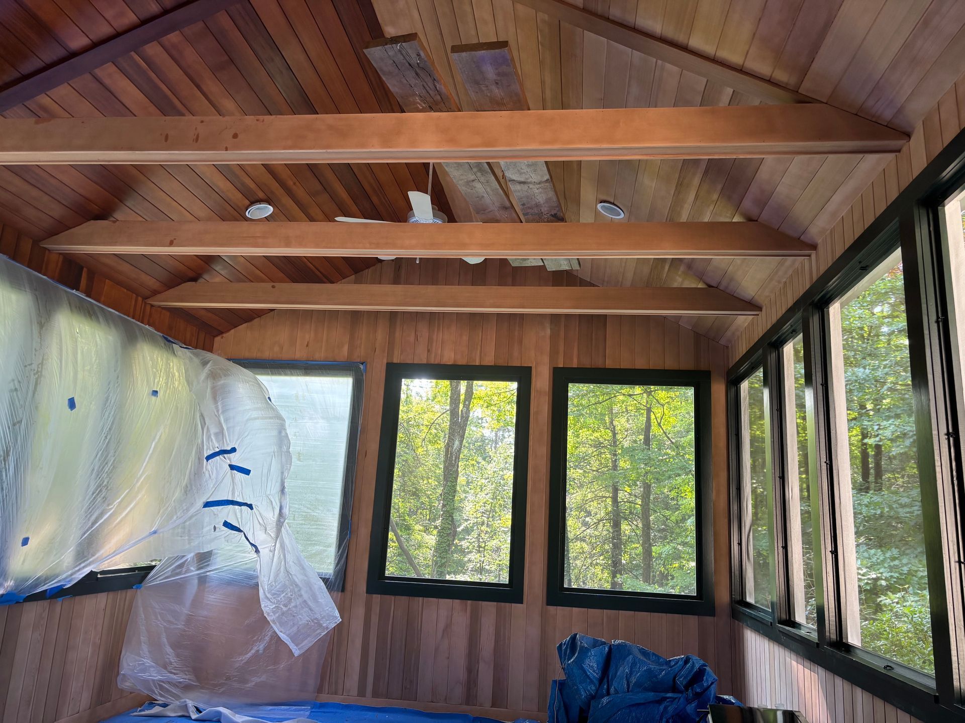 Wooden room interior with exposed beams, large windows, and plastic sheeting draped on the left.