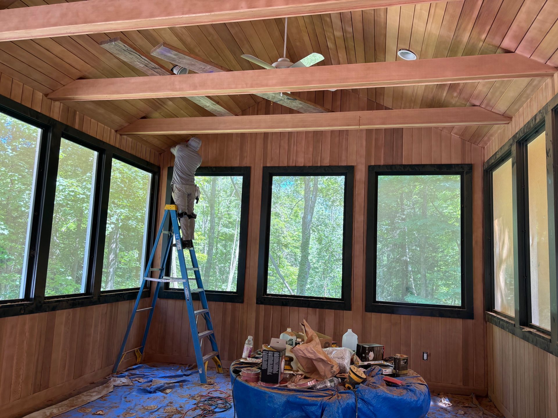 Wood-paneled room under construction with a blue ladder, exposed beams, and windows on three sides.