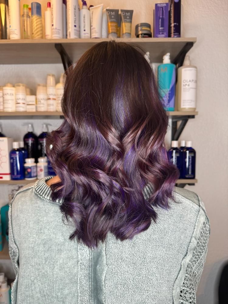 A woman with purple hair is standing in front of a shelf full of hair products.