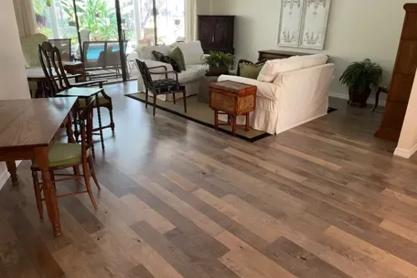 A sunlit living room featuring wood-plank floors, two beige sofas, patterned chairs, and a wooden table with chairs.